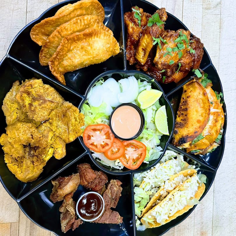 A black platter with various Tex-Mex dishes, including empanadas, wings, tacos, and plantains.